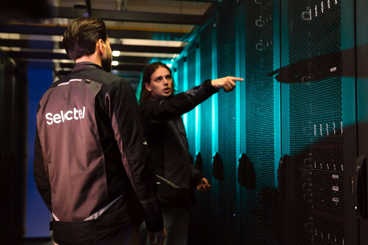 Специалисты Selectel что‑то оживленно обсуждают, стоя между рядов серверного оборудования.