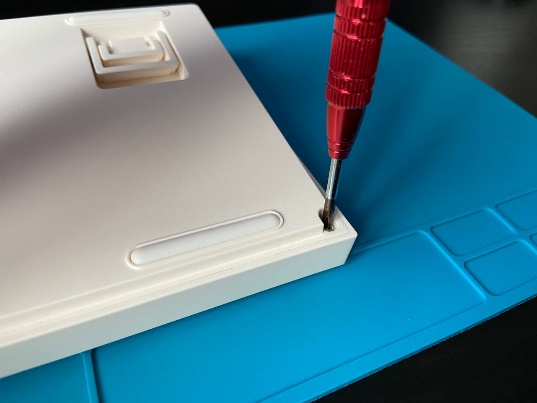 Откручивание крепежного винта в углу пластикового корпуса клавиатуры с помощью красной отвертки.